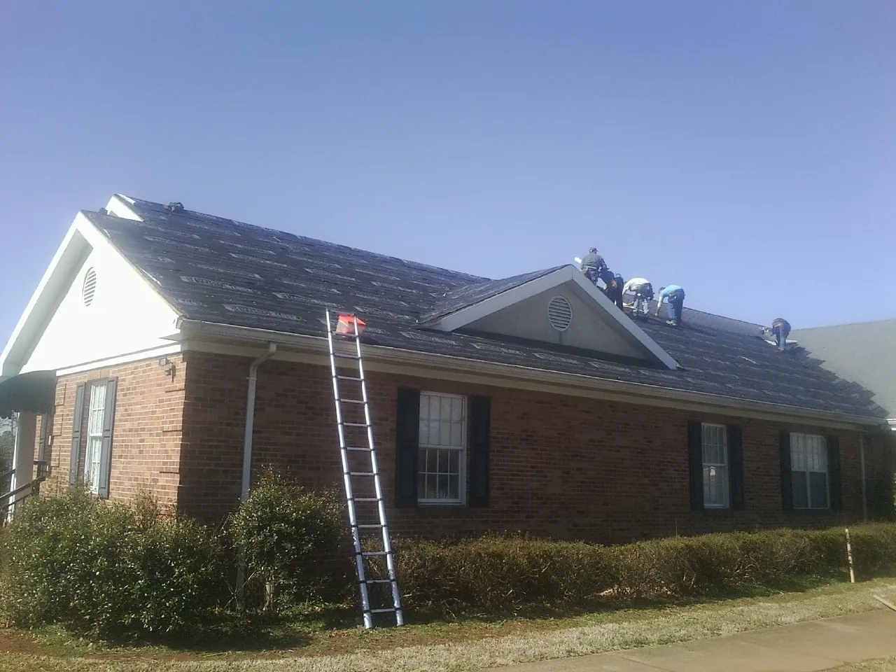 Crew tearing shingles off a brick ranch roof with a ladder set — Emergency Roof Repair work by our crew in Tea