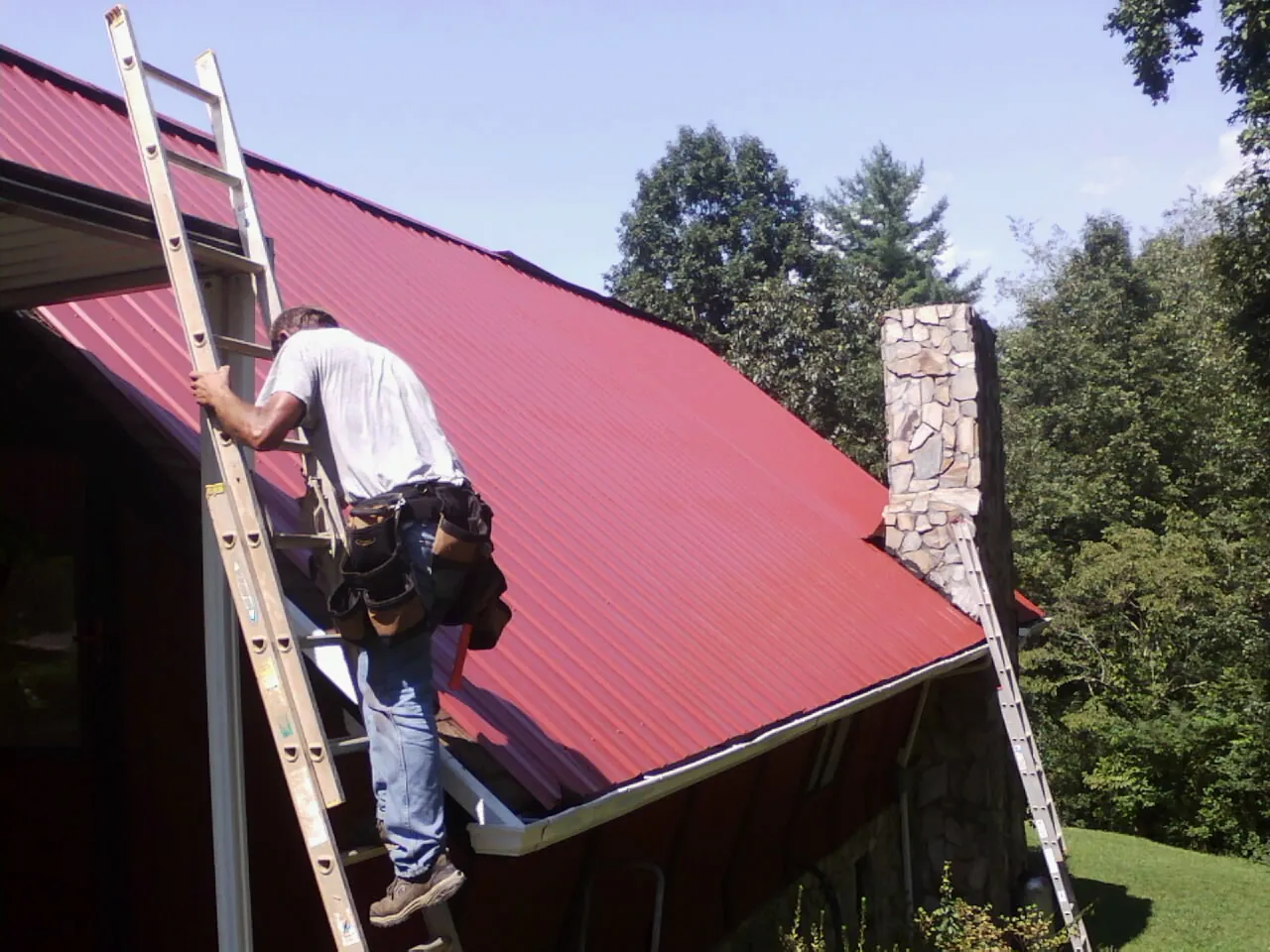 Roofer on a ladder beside a red metal roof and stone chimney — Soffit & Fascia Repair by Roofing Brothers in Tea